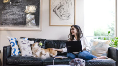 A woman with a laptop petting her dog on a sofa